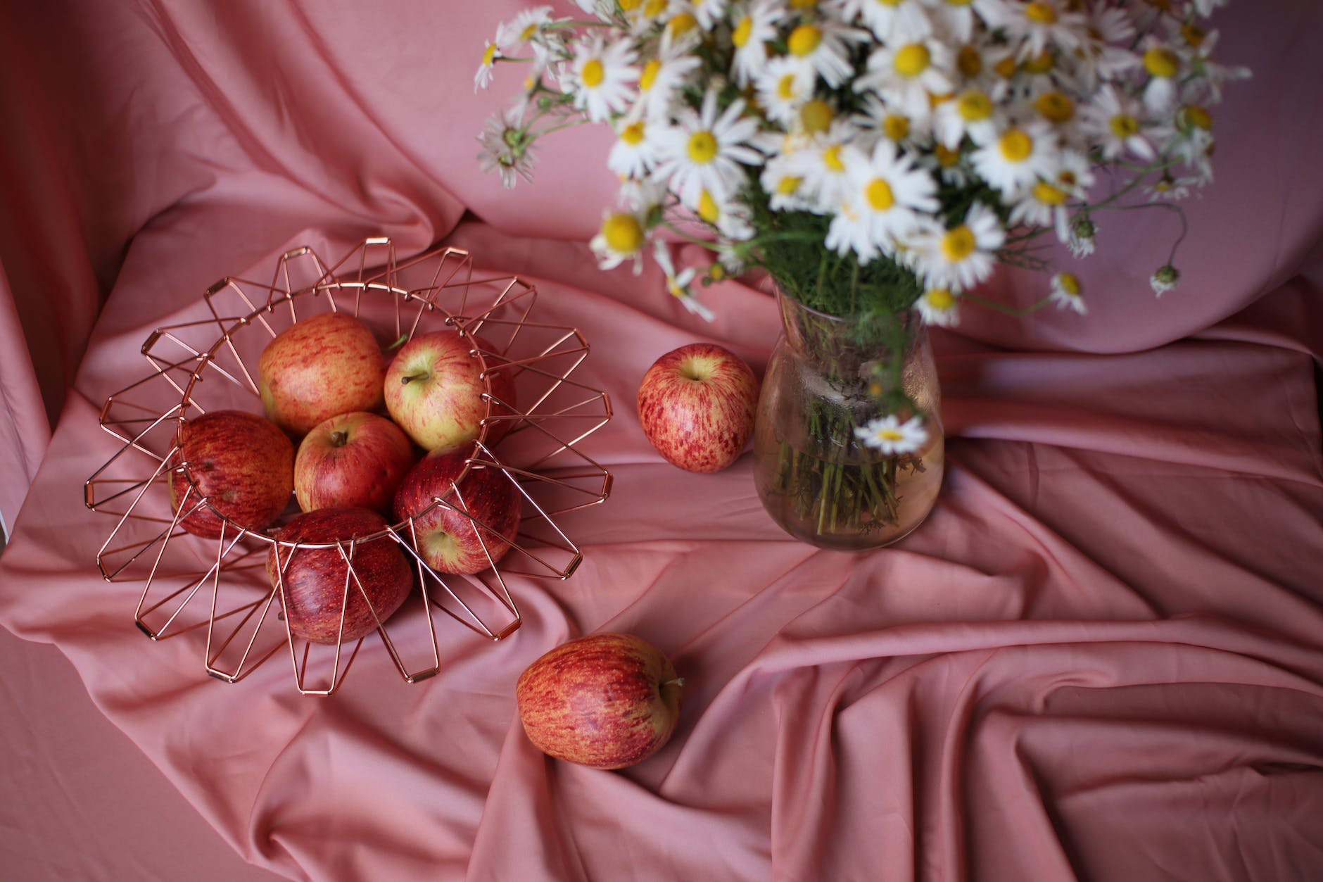red apples in metal basket and marigolds in glass vase on pink tablecloth