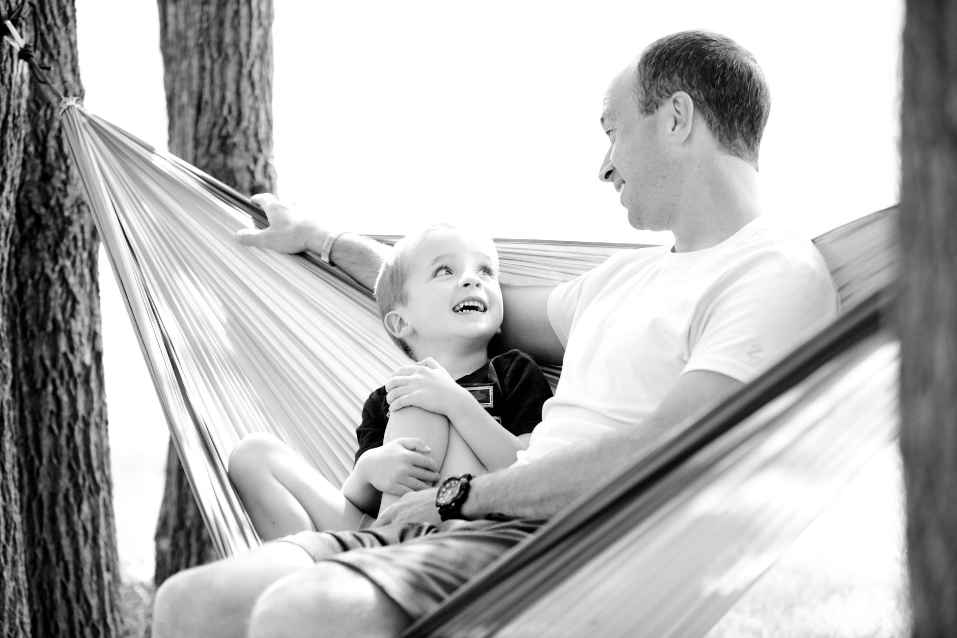 grayscale photo of man and child sitting on hammock
