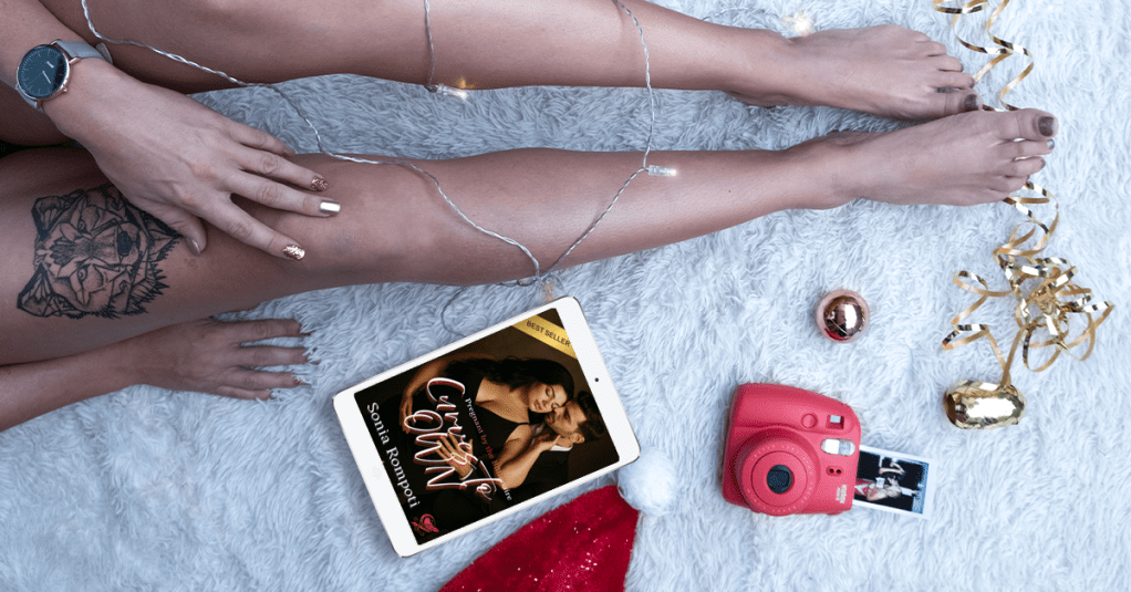 A close-up of a woman's legs with a tattoo, surrounded by holiday decorations, an e-reader displaying the book 'Curves to Own,' a camera, and festive ornaments.